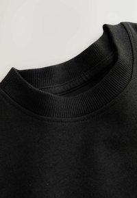 Sudadera negra con cuello redondo acanalado y tejido texturizado. El material suave tiene un aspecto liso sin patrones ni detalles.