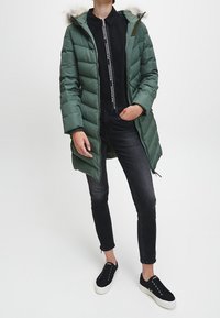 Abrigo de plumas verde con capucha con borde de piel, cierre de cremallera y diseño acolchado; combinado con jeans skinny negros y zapatillas negras.