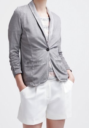 Osoba ubrana w jasnoszary żakiet nałożyła na pasekowany top oraz białe dopasowane szorty, z rękami w kieszeniach na jednolitym tle.