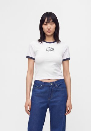 Femme portant un t-shirt court blanc Levi's avec bordure bleu marine et un jean bleu, debout devant un fond clair uni.