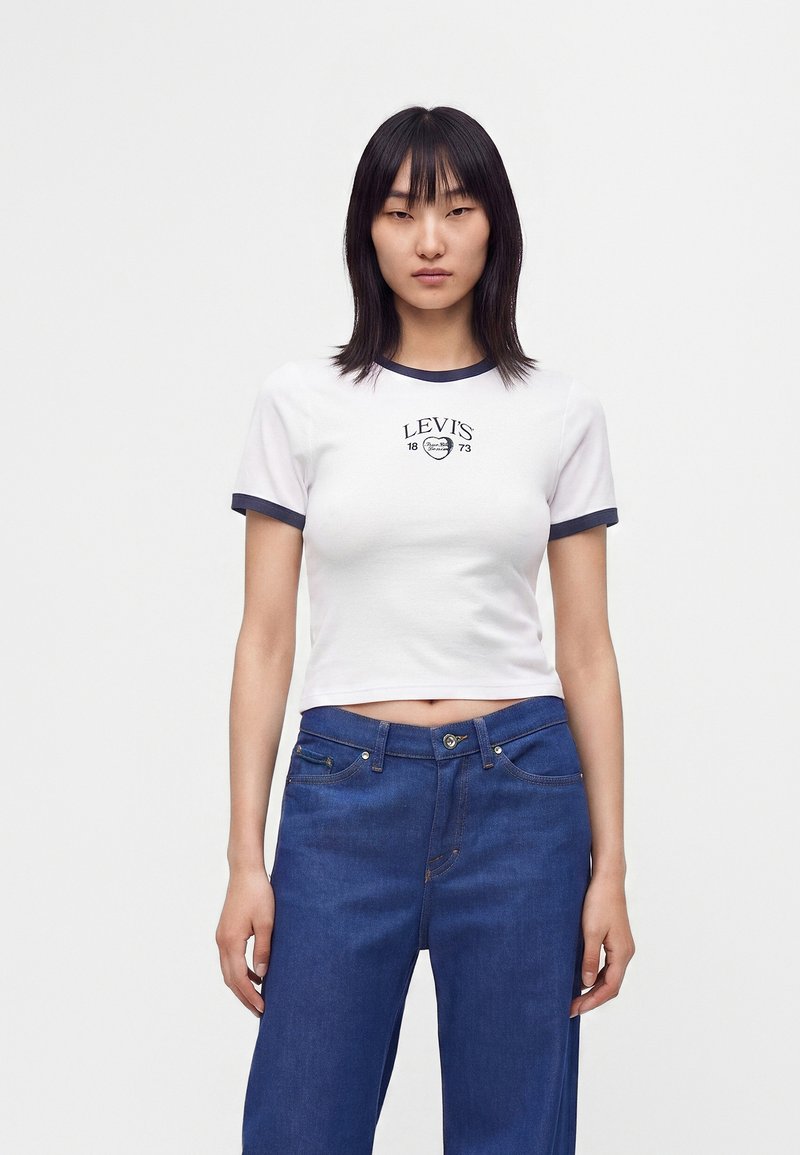 Femme portant un t-shirt court blanc Levi's avec bordure bleu marine et un jean bleu, debout devant un fond clair uni.