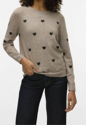 Femme portant un pull beige avec des motifs de cœurs noirs, associée à un jean bleu foncé, posant avec une main dans la poche.