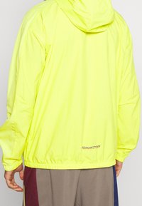 Gul letvægts vindjakke med hætte, elastiske manchetter og kant. Har "TOMMYSPORT" logoet på bagsiden. Glat tekstur, afslappet pasform.