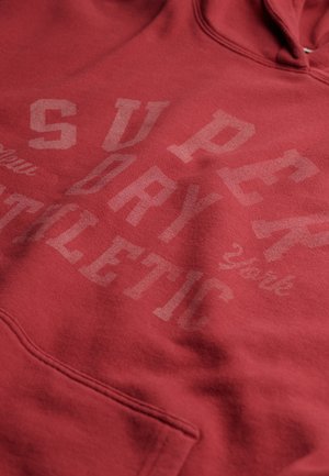 Czerwona bluza z kapturem o miękkiej fakturze, z przednią kieszenią oraz nadrukowanymi napisami w różnych rozmiarach: "SUPERDRY ATHLETIC" i "York."