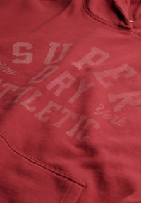 Rode hoodie van zachte stof, met een voorvak. Grafische elementen in wit en lichtgrijs tonen prominent "SUPERDRY ATHLETIC YORK".