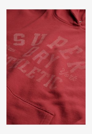 Felpa rossa con cappuccio realizzata in morbido tessuto, dotata di una tasca frontale. Le grafiche in bianco e grigio chiaro mostrano in modo evidente "SUPERDRY ATHLETIC YORK".