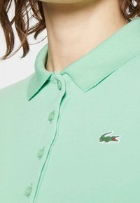 Camisa polo verde menta con cuello, que presenta una tapeta de botones y un pequeño logotipo bordado en el lado izquierdo del pecho. Textura de tela suave.