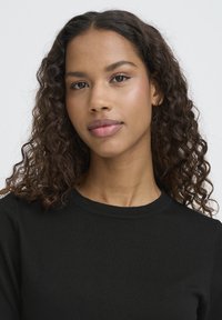 Pull noir côtelé avec un col rond. Le modèle a des cheveux ondulés brun foncé et un teint lisse. Design simple et épuré.