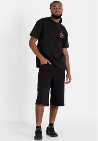Camiseta negra de manga corta con un pequeño gráfico rojo en el pecho, combinada con pantalones cortos negros hasta la rodilla y zapatillas negras.