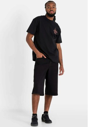 T-shirt nera a maniche corte con una piccola grafica rossa sul petto, abbinata a pantaloni corti neri fino al ginocchio e sneakers nere.