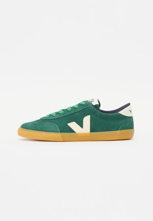 Sneaker en daim vert avec un logo crème, bout arrondi et semelle en caoutchouc texturé. Présente des lacets verts et un accent navy à l'arrière.