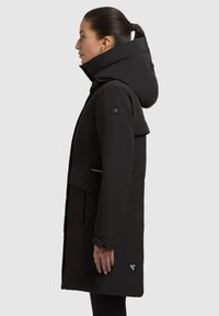 Kvinde med mørkt hår i hestehale, iført en lang sort vinterfrakke med hætte, står i profil mod en ensfarvet lys baggrund.