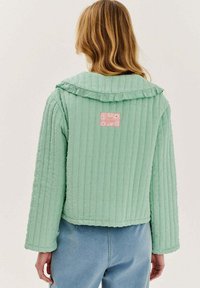 Svetle zelená prošívaná bunda s kapucí, s žebrovanou texturou a růžovou cedulkou zdobenou sedmikráskami. Má zkrácený střih a dlouhé rukávy.