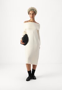 Λευκό πλεκτό φόρεμα σε στυλ σακακιού, μήκους μέχρι το γόνατο, με απαλή υφή. Μαύρ�η πλεκτή τσάντα χειρός και μαύρες κάλτσες έως τον αστράγαλο. Απλός σχεδιασμός, ελάχιστες λεπτομέρειες.