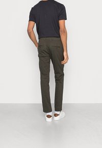 Donkergroene broek met een op maat gemaakte pasvorm, voorzien van achterzakken en subtiele stiksels. G gedragen met een zwart T-shirt en witte sneakers.