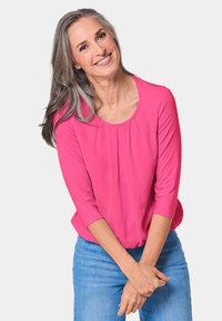 Fuchsia Bluse mit Dreiviertelärmeln, rundem Halsausschnitt und leichten Falten auf der Vorderseite, kombiniert mit hellblauen Jeans. Schlichtes, lässiges Design.