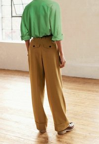 Groen, lang mouwen shirt gecombineerd met hooggesneden, wijde mosterdkleurige broek met achterzakken en donkere knopen, gedragen met bruine schoenen.