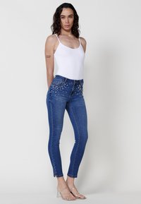 High-waisted blue skinny jeans with denim texture, embellished with pearl-like accents along the front pockets. Paired with a white tank top.