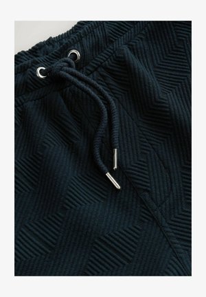 Mørke joggers med struktur, der har et herringbone-mønster, elastisk talje og snor med metalspidser.