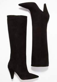 Une paire de bottes noires en daim, hautes, avec des orteils pointus et des talons coniques, une botte debout et l'autre couchée sur le côté.