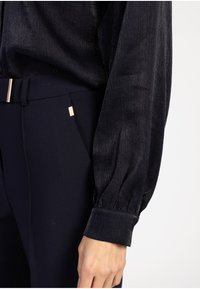 Close-up van een persoon die een donkere, getextureerde blouse met een manchet-sleeve en een op maat gemaakte marineblauwe broek met zijzak en riemdetail draagt.