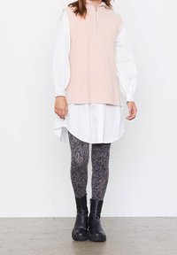 Helles rosa ärmelloses Strickoberteil mit hohem Kragen, übereinander getragen mit einem weißen Hemd mit Knöpfen, kombiniert mit schwarzer und grauer gemusterter Leggings und schwarzen Stiefeln.
