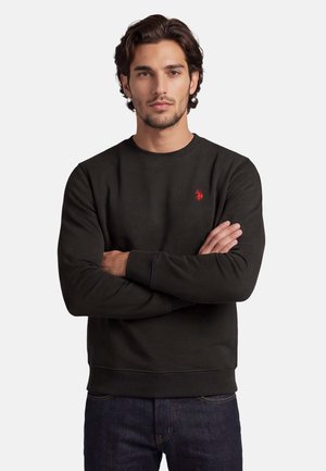 Man with dark hair wearing black long-sleeve sweatshirt with red logo, arms crossed, standing against plain light background.