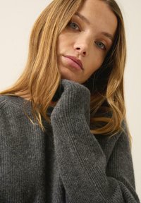 Harmaa ribattua neulepuseroa, jossa on rento istuvuus ja pitkät hihat, leveä pääntie ja pehmeä tekstuuri. Ei näkyviä kuvioita tai koristeita.