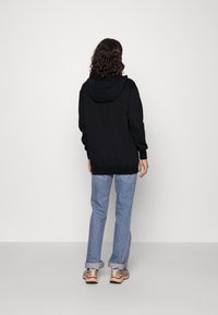 Sudadera negra con capucha y mangas largas, llevada sobre unos jeans azul claro y zapatillas. El material parece suave y tiene un corte relajado.