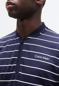 Κοντινό πλάνο ενός ατόμου που φοράει μπλε ναυτικό και άσπρη ριγωτή μπλούζα με φερμουάρ Calvin Klein και κοντό γιακά.