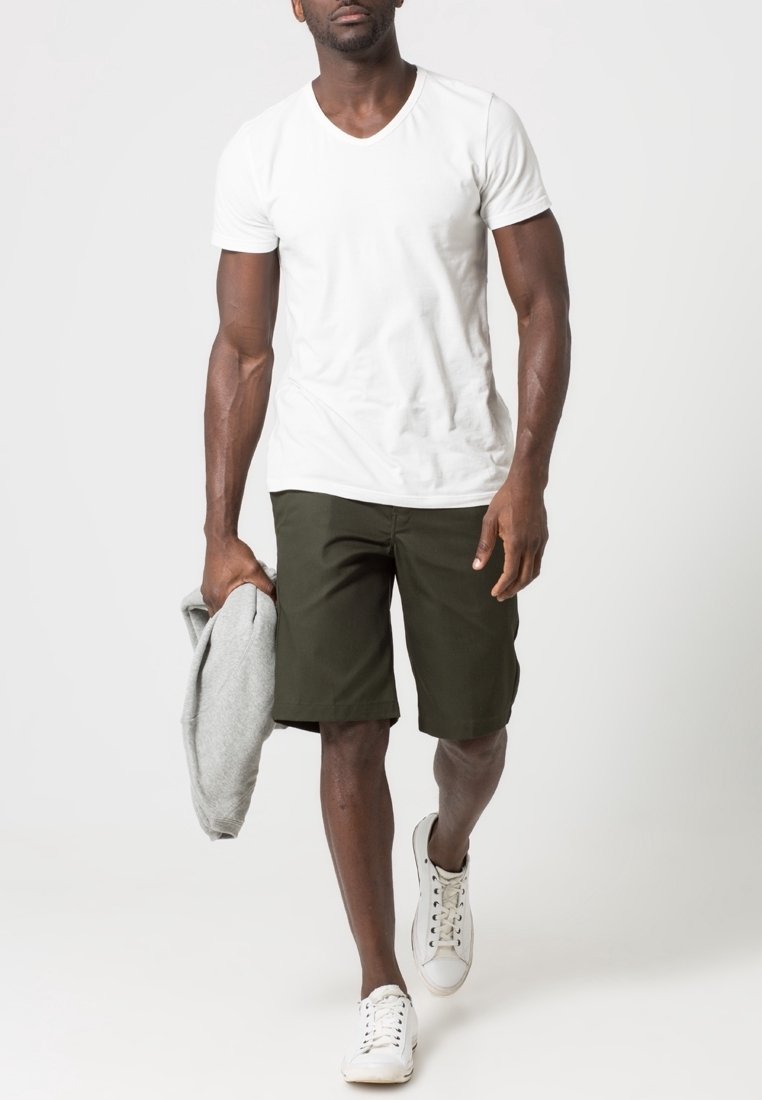 Homme marchant portant un t-shirt blanc à manches courtes, un short vert foncé, des baskets blanches, tenant un sweat gris plié dans une main.