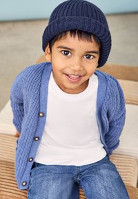 Cardigan tricotat de un albastru deschis, cu nasturi, peste un tricou alb, purtat cu blugi. Băiatul poartă o bustieră căptușită de un albastru închis.