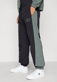 Juodos ir pilkos sportinės kelnės su elastinėmis pūtimis, šoninėmis juostelėmis ir logotipu. Pagamintos iš lengvos medžiagos, su raištelių juosmeniu.