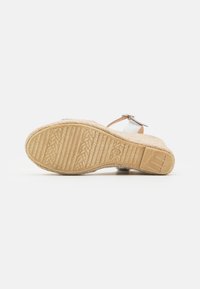 Sandale compensée avec une semelle beige texturée, des lanières argentées et une accentuation marron. Le design inclut une boucle à la cheville pour un maintien sécurisé.