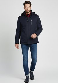 Hombre vestido con una chaqueta con capucha azul marino sobre una camisa de cuadros rojos, jeans azul oscuro ajustados y botas negras, caminando hacia adelante contra un fondo liso.