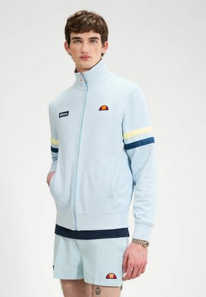 Jeune homme portant une veste zippée bleu clair et un short assorti avec des logos de marque et des rayures sur les manches, debout devant un fond uni.