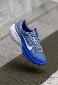 Pelēki un zili sporta apavi ar teksturētu virsu, balts Nike zīmols un elpojoša acs. Mīkstināta zole ar noapaļotu pirkstu dizainu.