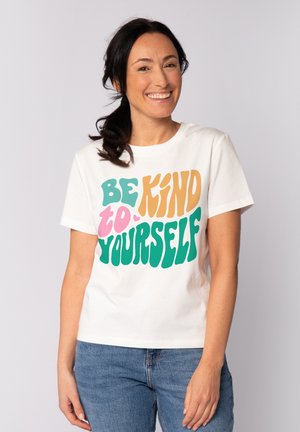 Frau lächelt, trägt ein weißes T-Shirt mit buntem Schriftzug "Sei nett zu dir selbst", kombiniert mit blauen Jeans, stehend vor einem einfarbigen hellen Hintergrund.