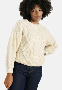 Krémový sveter s pleteným vzorom, s rebrovaným lemom a manžetami. Má textúrovaný vzor a voľný strih, v kombinácii s tmavými džínsami.
