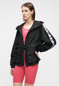 Γυναίκα που φοράει μαύρο μπουφάν με φερμουάρ και λευκή ρίγα με λογότυπο στα μανίκια, πάνω από ροζ αθλητικό σουτιέν και ταιριαστό ροζ ποδηλατικό σορτς.