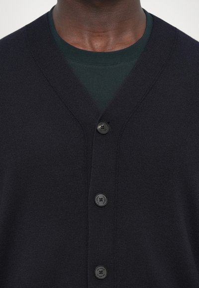 Cardigan de marin cu decolteu în V, butoane negre și textură moale. Purtați peste un tricou verde închis, evidențiind un aspect stratificat.