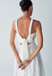 Weißes Kleid mit tiefem Rückenausschnitt, elastischen Trägern und einer gebundenen Schleife in der Taille. Der Stoff ist glatt mit leichtem Glanz und einer lockeren Silhouette.