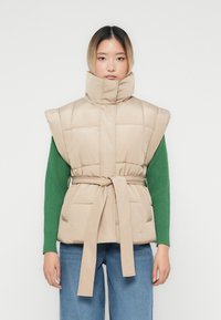 Ženska, oblečena v bež podloženo brezrokavnik z pasom, visokim ovratnikom, zelenim puloverjem in modrimi jeans hlačami, stoji ob beli steni.