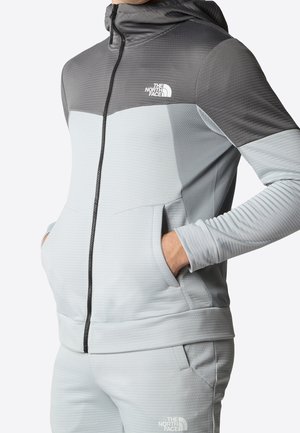 Homme portant un sweat à capuche zippé gris clair et gris foncé assorti avec des logos The North Face, les mains dans les poches du sweat.