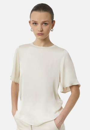 Haut en crème soyeux avec des manches courtes à volants et un col rond. Texture lisse et coupe décontractée, associé à un pantalon de teintes neutres.