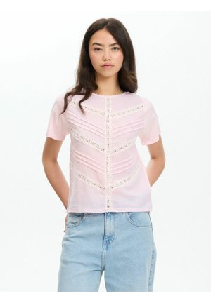 Joven mujer que lleva una blusa de manga corta de color rosa claro con detalles de encaje y vaqueros de tiro alto azul claro, de pie con las manos detrás de la espalda.