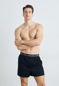 Čierne boxerky s bielym pásom s logom "KARL LAGERFELD". Materiál vyzerá mäkko, má jeden predný gombík a pohodlný strih.