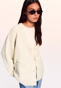 Cardigan oversize color crema con polsini a coste e due tasche frontali, indossato sopra una camicia chiara, abbinato a occhiali da sole rotondi neri.