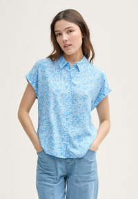Blusa azzurra a maniche corte con piccolo motivo floreale bianco, chiusura con bottoni e vestibilità ampia. Abbinata a pantaloni in denim azzurro.