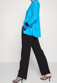 Une personne marche dans un blaser bleu saisissant associé à un pantalon noir et des talons noirs à lanières, mettant en avant un look de mode contemporain.